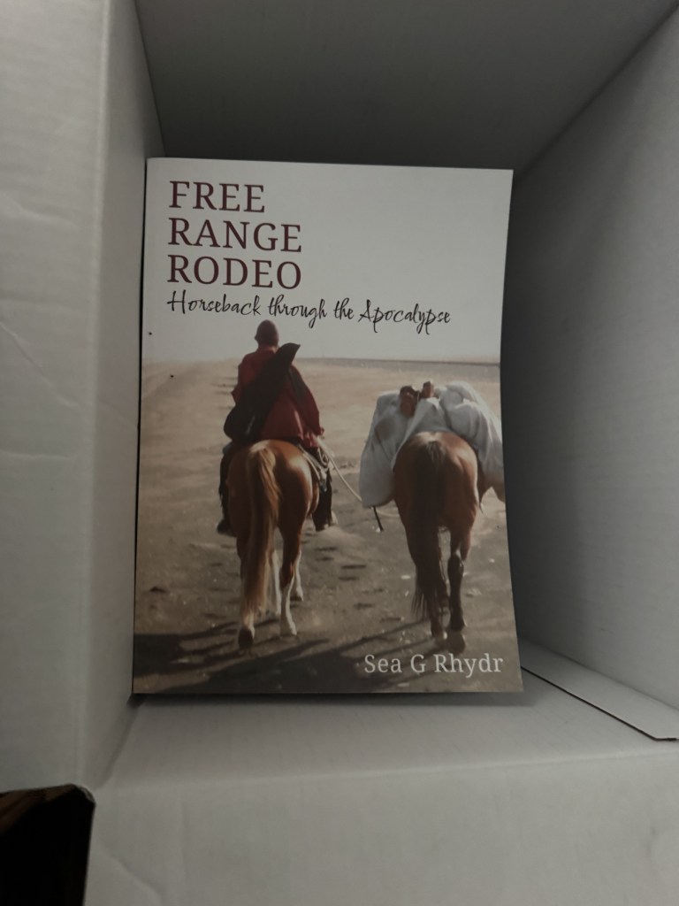 A book titled 'Free Range Rodeo: Horseback through the Apocalypse' by Sea G Rhydr, placed inside a box with a cover image showing two riders on horses.