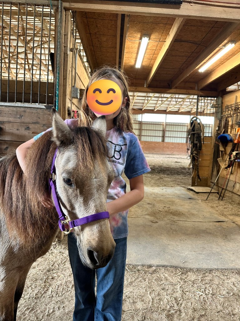 A child stands beside a small horse in a barn, gently holding its head. The child is wearing a colorful tie-dye shirt and jeans, with an emoji covering their face.