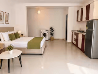 A modern studio apartment featuring a bed with green accent pillows, a kitchenette with cabinets, and a washing machine. A small round table with a plant is in the foreground.