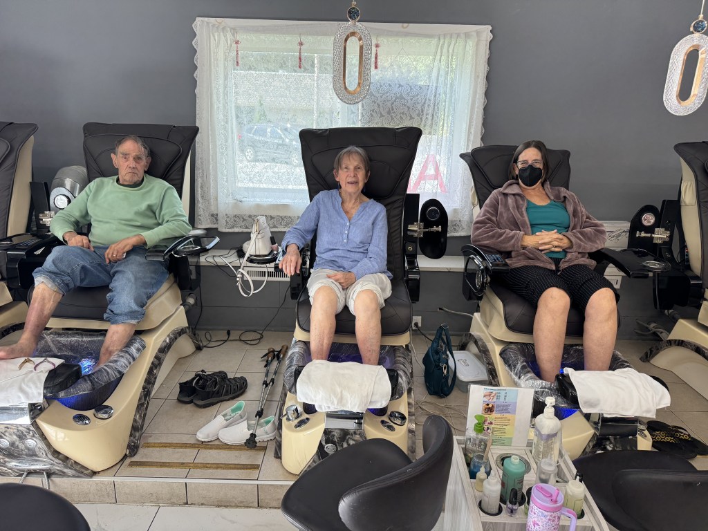 Three individuals relaxing in massage chairs at a nail salon, with various nail care products visible in the foreground.