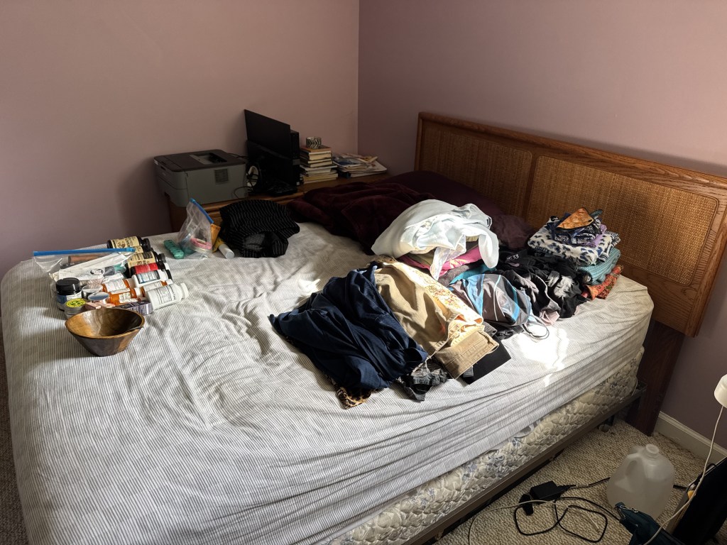 A messy bedroom with a bed covered in folded clothes, a bowl, and various items arranged around a printer and a stack of books on a nearby table.