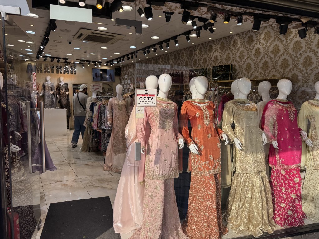 Storefront displaying a variety of elegant traditional dresses on mannequins, with a decorative interior featuring intricate wallpaper.