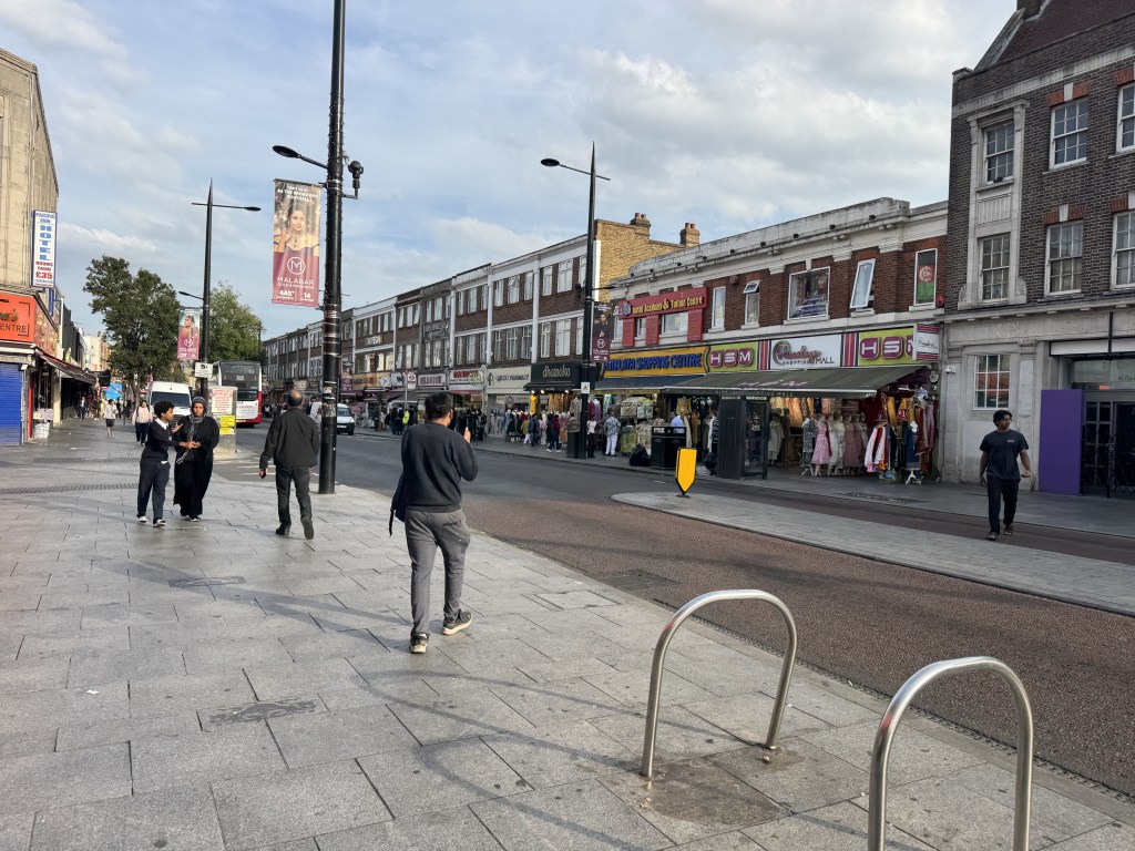 A busy urban street scene with people walking along the pavement, shops with colorful storefronts on either side, and a bus in the background. The sky is partly cloudy.
