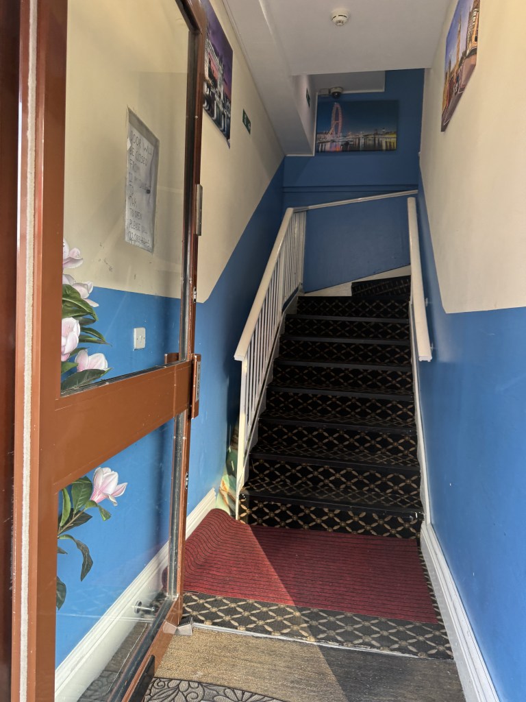 Interior view of a stairway leading upwards, featuring blue walls and decorative posters. A double door entrance is visible with a floral design on the left side. The stairs are carpeted with a patterned runner, and the area is well-lit.