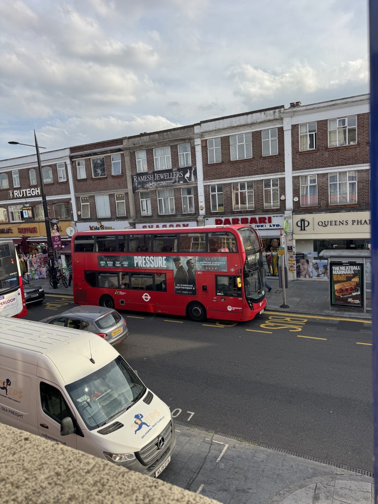 A red double-decker bus with advertisements stops on a busy urban street, surrounded by shops and parked vehicles. In the background, there are various storefronts and pedestrians.