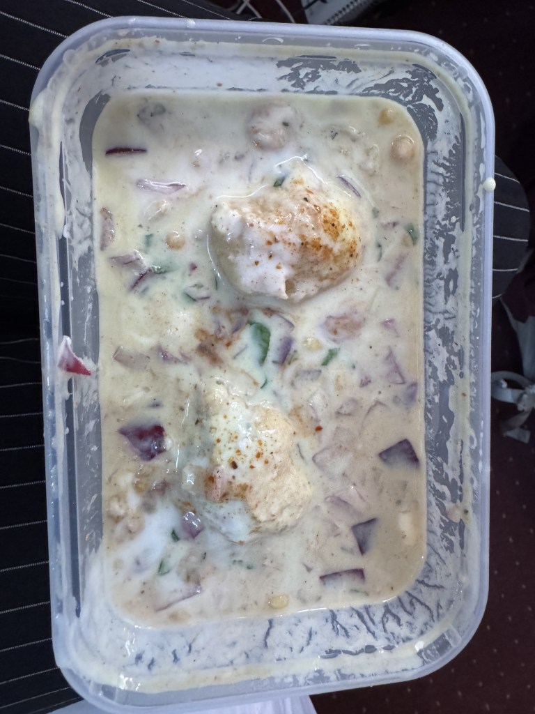 A serving of yogurt-based dish featuring chopped vegetables and soft boiled eggs, topped with spices, served in a plastic container.