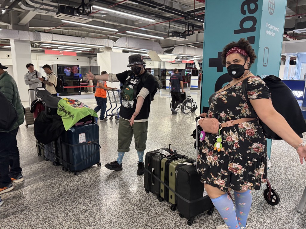 Two travelers at an airport check-in area posing with their luggage. One person is wearing a black T-shirt and mask, with colorful luggage and a green shirt draped over it. The other is wearing a floral dress and has a backpack.